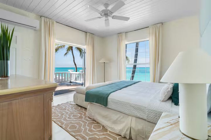 Sunlit beachfront bedroom with white linens and teal throw, balcony and windows framing turquoise ocean and swaying palm trees, coastal decor and ceiling fan.