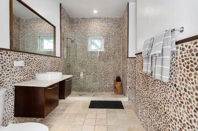 Spa-like modern bathroom with pebble-tile walls, glass walk-in shower, floating dark-wood vanity with white vessel sink, small window, striped gray towels and beige tile floor.