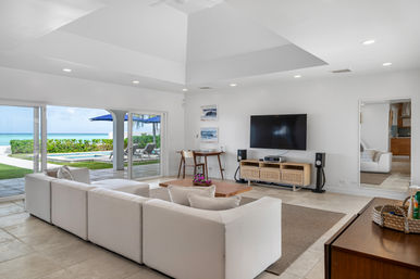 Bright modern beachfront living room with white modular sectional, wood coffee table and wall-mounted TV; sliding glass doors open to a patio with pool, lounge chairs and turquoise ocean beyond.