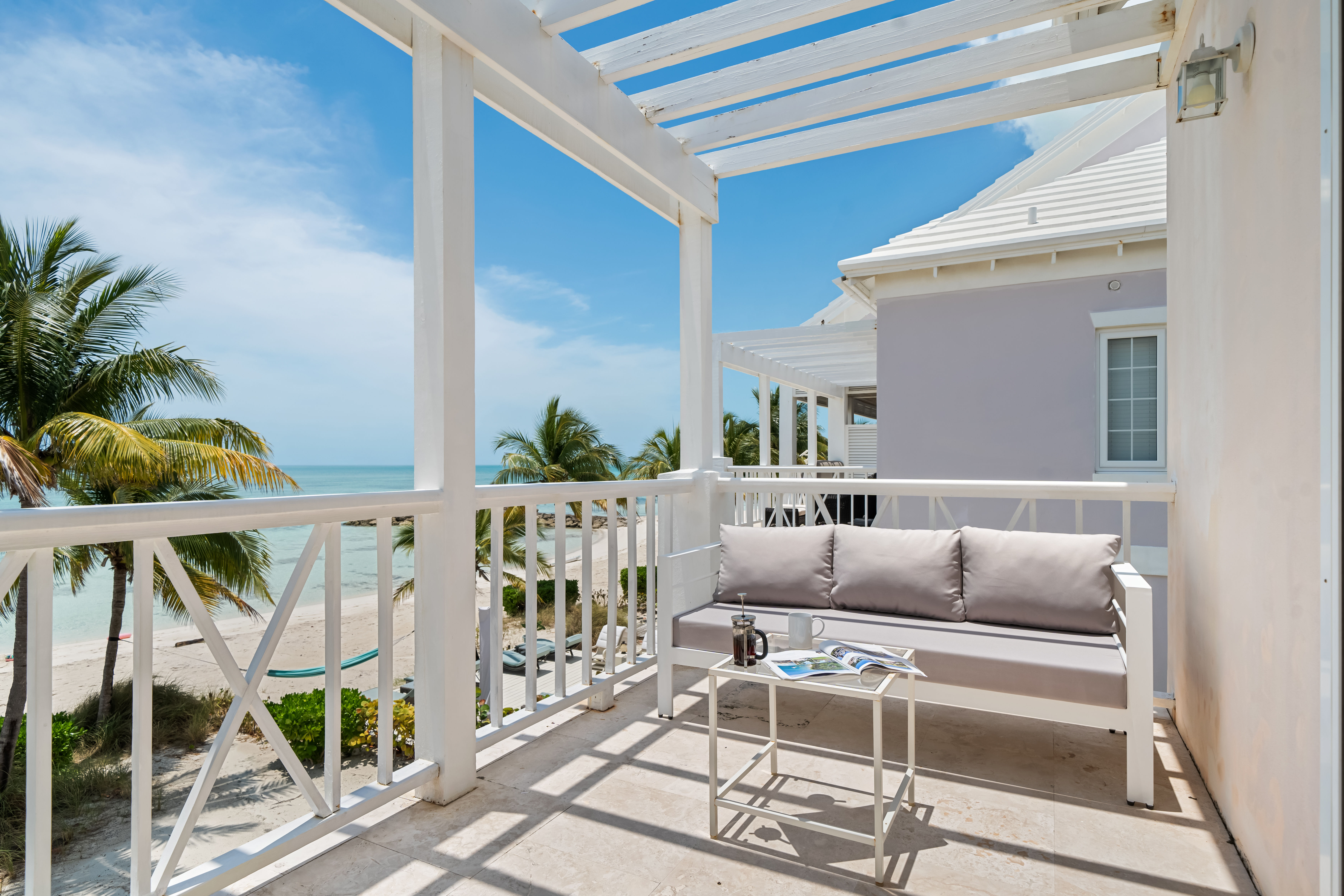 Sunny beachfront balcony with white wooden pergola, cushioned outdoor sofa and small table overlooking turquoise ocean, sandy beach and swaying palm trees — perfect spot for morning coffee.