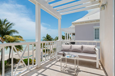 Sunny beachfront balcony with white wooden pergola, cushioned outdoor sofa and small table overlooking turquoise ocean, sandy beach and swaying palm trees — perfect spot for morning coffee.