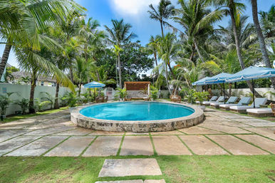 Round turquoise swimming pool at a tropical resort surrounded by palm trees, blue umbrellas, cushioned sun loungers, a wooden cabana and stone patio under a sunny sky