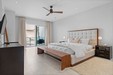 Sunlit modern condo master bedroom with tufted upholstered king bed, wooden bench at the foot, neutral bedding, ceiling fan, bedside tables with globe lamps, wall-mounted TV on a dresser and sliding glass doors opening to a small balcony with outdoor seating.