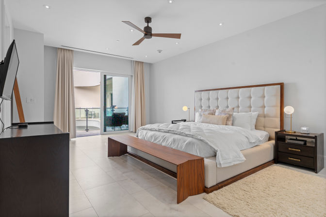 Sunlit modern condo master bedroom with tufted upholstered king bed, wooden bench at the foot, neutral bedding, ceiling fan, bedside tables with globe lamps, wall-mounted TV on a dresser and sliding glass doors opening to a small balcony with outdoor seating.