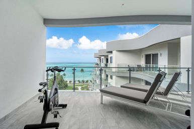 Modern oceanfront balcony with an exercise bike and two gray lounge chairs behind a glass railing, overlooking turquoise tropical sea and palm trees.
