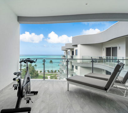 Modern oceanfront balcony with an exercise bike and two gray lounge chairs behind a glass railing, overlooking turquoise tropical sea and palm trees.