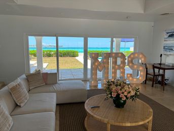 Beachfront living room with white sectional sofa, sliding glass doors framing turquoise ocean and patio, illuminated marquee letters and a vase of pink roses on a wicker coffee table.