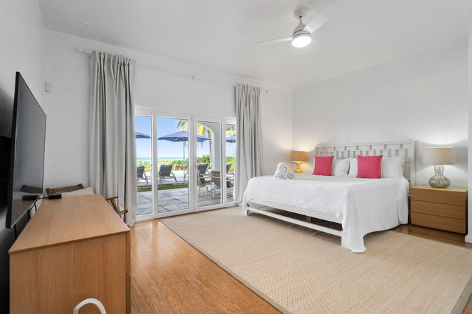 Bright beachfront bedroom with king bed in white linens and pink accent pillows, wood nightstands and lamps, sliding glass doors opening to a patio with lounge chairs, blue umbrellas, palm trees and ocean view.