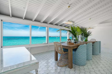 Sunlit oceanfront coastal dining room with panoramic turquoise sea view through large windows, wooden table with rattan chair, pale blue wavy-upholstered stools, tropical palm centerpieces, white beamed ceiling and hex tile floor