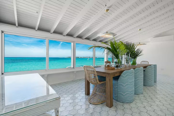 Sunlit oceanfront coastal dining room with panoramic turquoise sea view through large windows, wooden table with rattan chair, pale blue wavy-upholstered stools, tropical palm centerpieces, white beamed ceiling and hex tile floor