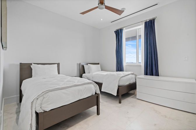 Bright minimalist twin bedroom with two single beds and white linens, dark wood frames, navy curtains at a window showing blue sky, white low dresser, ceiling fan and light tile floor — modern, clean aesthetic.