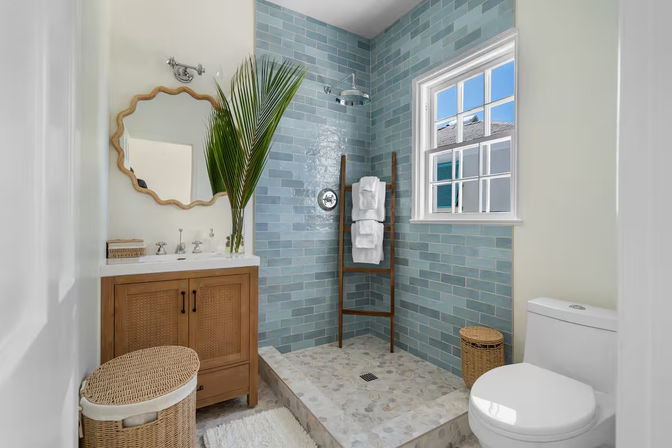 Bright coastal-style bathroom with blue subway-tile walk-in shower, wooden ladder towel rack, wood vanity, woven wicker baskets, palm frond accent and sunlit window.
