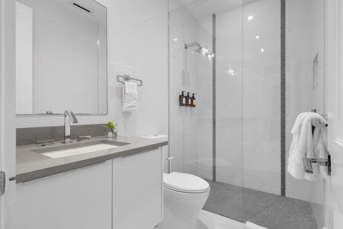 Sleek modern bathroom with gray stone vanity and undermount sink, large mirror, frameless glass walk-in shower with wall-mounted soap dispensers, toilet and neatly folded towels — spa-like design.