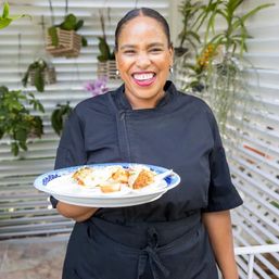 Smiling chef in a black uniform holding a blue-patterned plate of bite-sized appetizers on a sunny garden patio with hanging plants