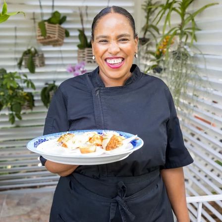 Smiling chef in a black uniform holding a blue-patterned plate of bite-sized appetizers on a sunny garden patio with hanging plants