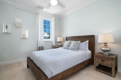 Bright coastal-style master bedroom with a wooden platform bed, light blue-gray walls, patterned pillows, white curtains framing a window with palm-tree view, matching wooden nightstands, metallic lamps and light hardwood floors.