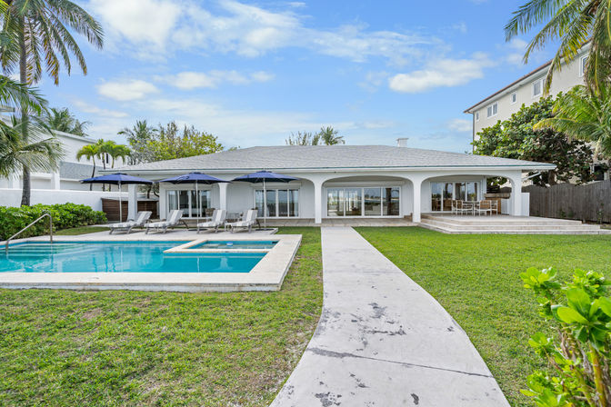 Waterfront single-story vacation home with pool, lounge chairs and umbrellas, patio dining and palm trees