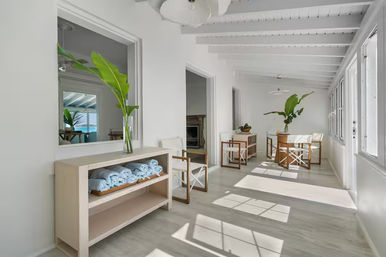 Sunlit coastal sunroom hallway with white beamed ceiling, light wood shelving holding rolled blue towels, folding director-style chairs, tropical banana-leaf plants and large windows with ocean view.