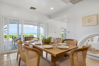 Bright coastal dining room with wooden table set for six, rattan-cane chairs and sliding glass doors opening to a beachfront patio and turquoise ocean view.