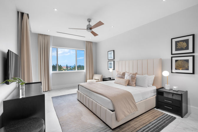 Bright modern ocean-view master bedroom with a beige upholstered king bed, neutral linens and throw, ceiling fan, black nightstands with globe lamps, large window overlooking treetops and blue horizon