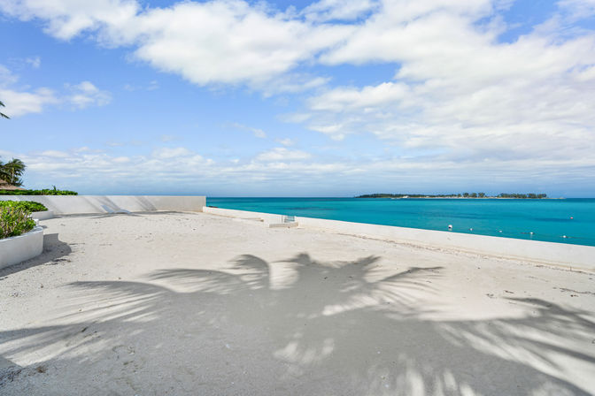 Tropical oceanfront terrace with white sand and palm-leaf shadows, turquoise sea and distant island on the horizon under a blue sky