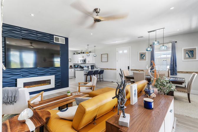 Open-concept modern coastal living room and kitchen with mustard-yellow leather sofa, navy textured TV accent wall and electric fireplace, wall-mounted flat-screen, white island with bar stools, blue pendant lights and sunny dining area.