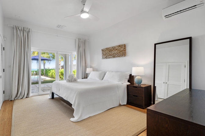 Bright coastal bedroom with king bed in white linens, ceiling fan and full-length mirror; sliding glass doors open to a patio with palm trees, umbrella and a glimpse of the ocean — minimalist tropical retreat