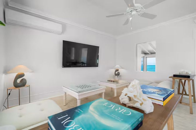 Bright coastal beach-house living room with white walls and ceiling fan, wall-mounted TV, two white game tables with chessboard, modern sculptural lamps, ocean view through a window, and blue coffee-table books with coral decor.