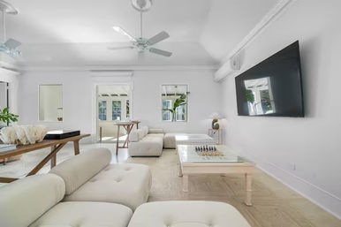 Bright airy minimalist living room with white modular sectional, glass coffee table with chess set, wall-mounted flat-screen TV, ceiling fans, wooden console table and sunlit windows
