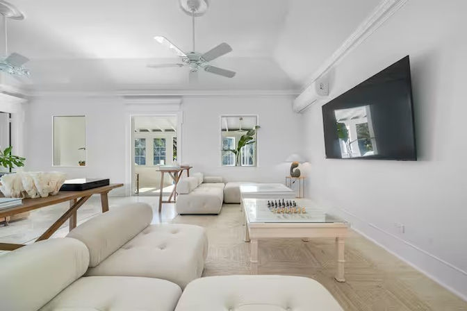 Bright airy minimalist living room with white modular sectional, glass coffee table with chess set, wall-mounted flat-screen TV, ceiling fans, wooden console table and sunlit windows
