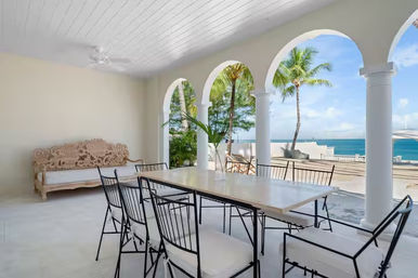 Oceanfront covered veranda with arched columns and a long dining table surrounded by wrought-iron chairs, carved wooden bench, palm trees and turquoise sea views.