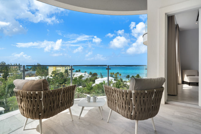 Oceanfront balcony with two woven lounge chairs and a small white table with coffee mugs, glass railing framing turquoise sea, palm trees and blue sky.