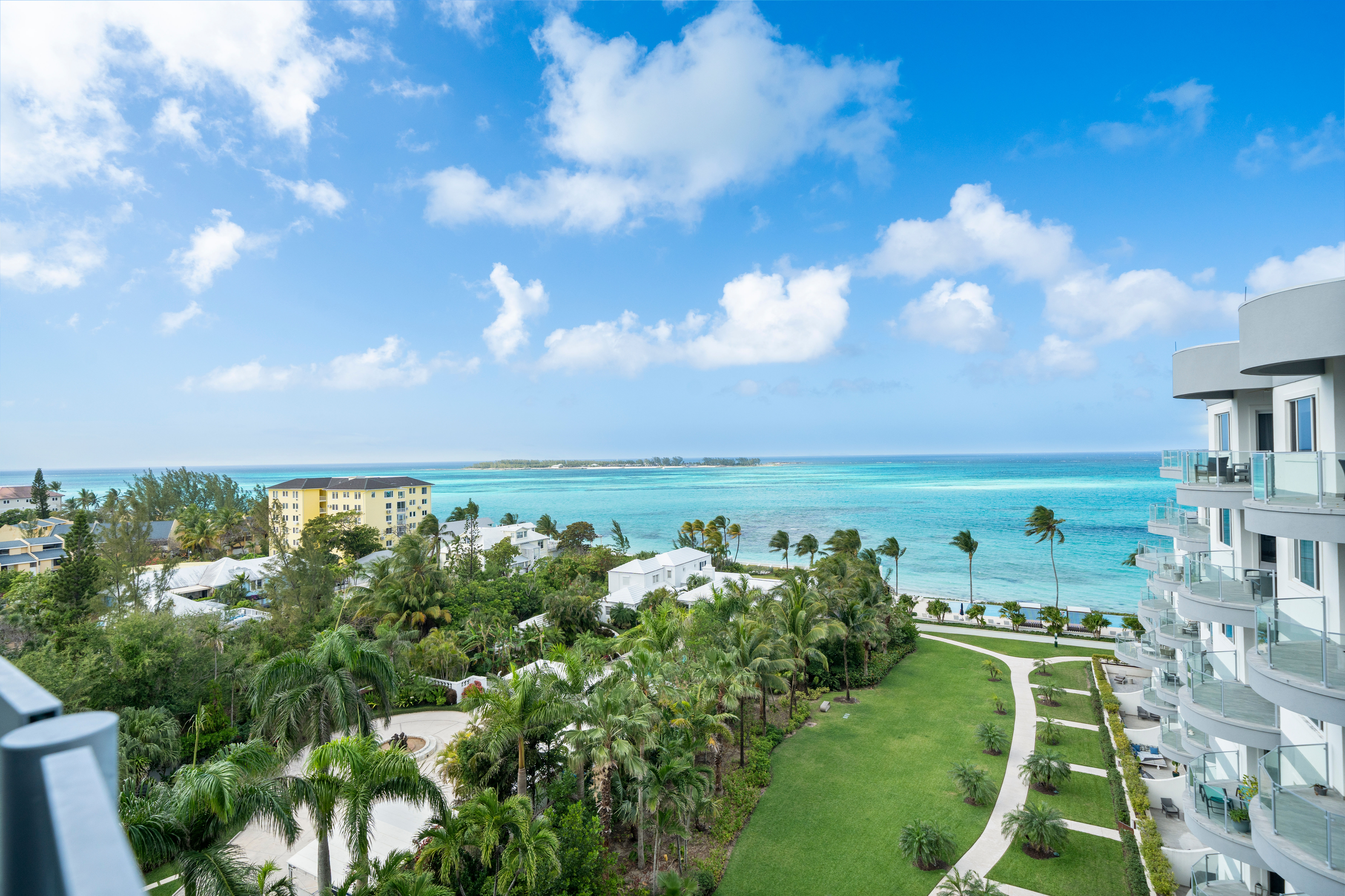 Sunny oceanfront view from a modern condo balcony: turquoise tropical sea, palm‑lined landscaped lawn and winding beachfront pathway with low-rise coastal buildings and a distant island on the horizon.