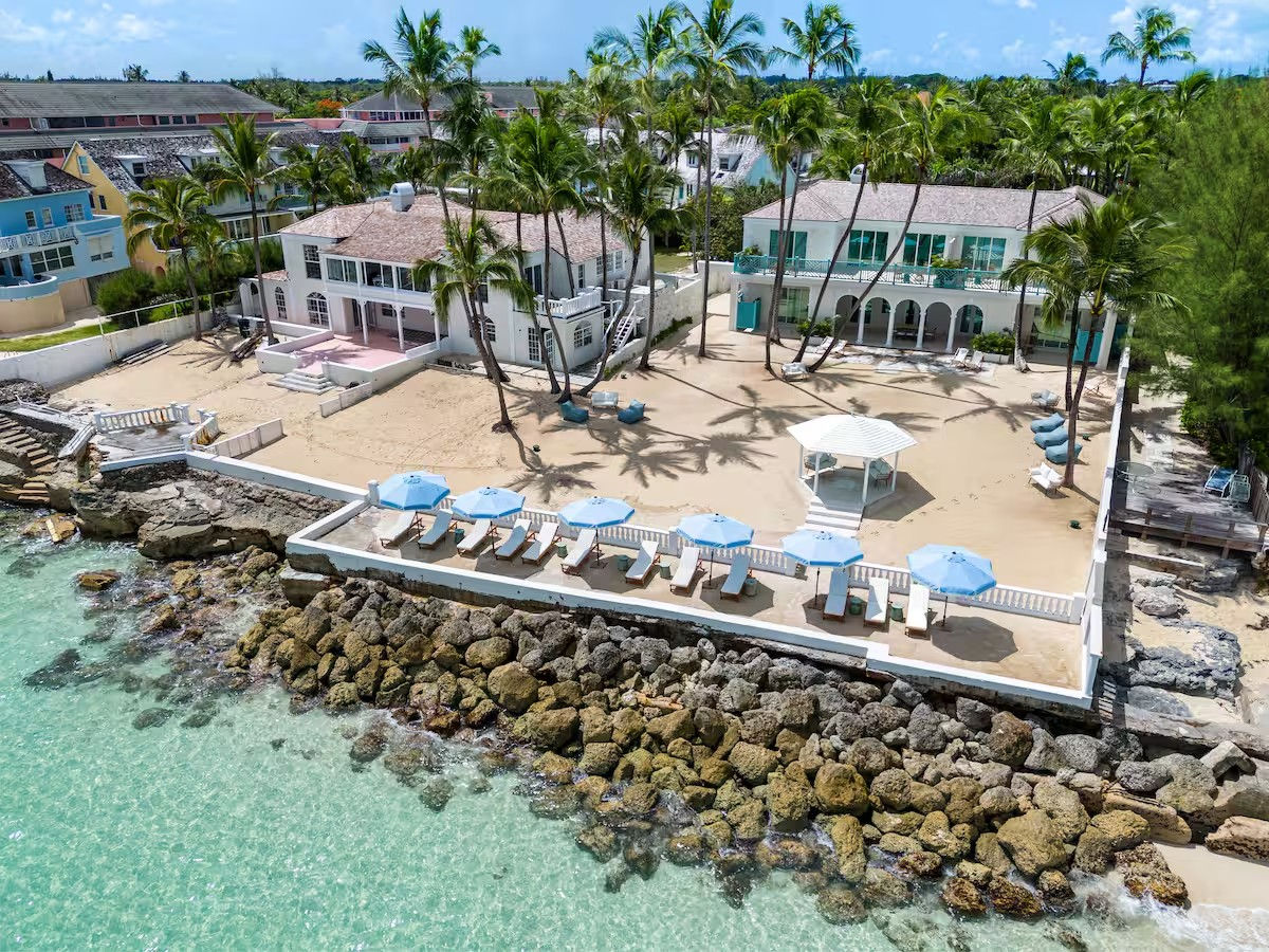 Aerial view of sun‑soaked tropical beachfront villas with palm trees, sandy courtyard and white gazebo, blue-umbrella lounge chairs on a seawall above turquoise water and rocky shoreline.