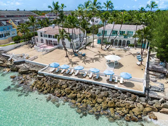 Aerial view of sun‑soaked tropical beachfront villas with palm trees, sandy courtyard and white gazebo, blue-umbrella lounge chairs on a seawall above turquoise water and rocky shoreline.