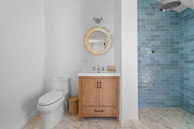 Bright coastal-style bathroom with wooden vanity and white sink, decorative round gold mirror, white toilet and wicker basket, blue subway-tiled walk-in shower with rainfall showerhead and hexagon mosaic floor tiles