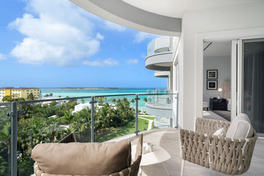 Oceanfront balcony with woven lounge chairs and small table overlooking turquoise tropical sea, palm trees, and curved modern balconies under a blue sky.