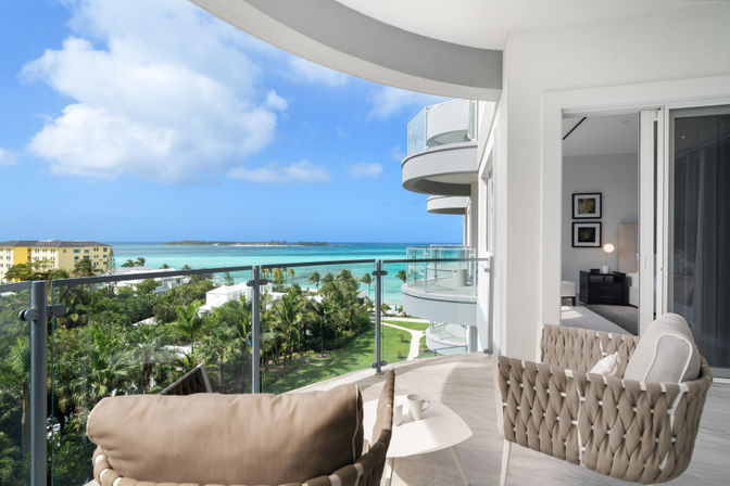 Oceanfront balcony with woven lounge chairs and small table overlooking turquoise tropical sea, palm trees, and curved modern balconies under a blue sky.