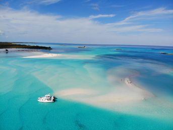 Aerial view of a picture-perfect tropical lagoon with turquoise, crystal-clear water, a white sandbar with a few people, and an anchored yacht under a bright blue sky.