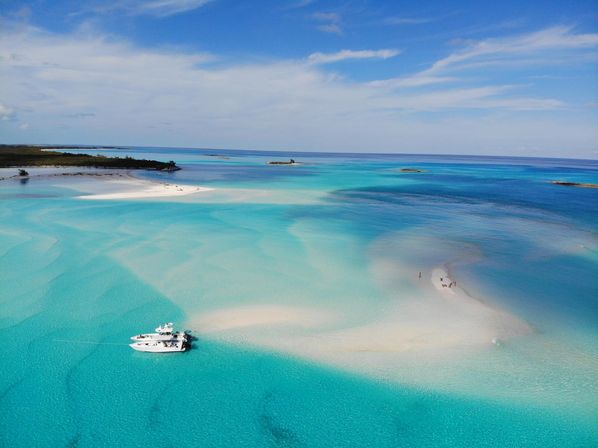 Aerial view of a picture-perfect tropical lagoon with turquoise, crystal-clear water, a white sandbar with a few people, and an anchored yacht under a bright blue sky.