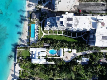 Aerial view of a beachfront property with turquoise water and white-sand beach, palm trees, a long oceanfront swimming pool and a smaller central pool among white buildings and landscaped lawns.