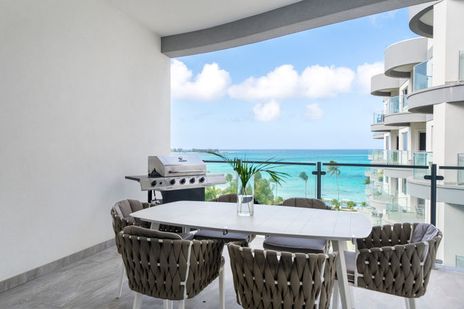 Oceanfront condo balcony with white dining table, woven chairs, gas grill, palm trees and turquoise sea view.