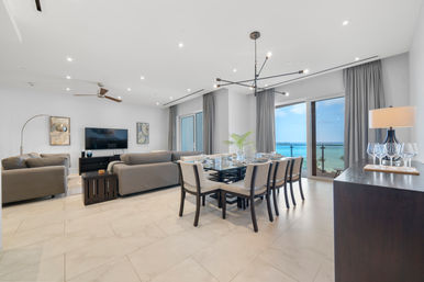 Bright modern beachfront condo living and dining area with gray sofas, glass dining table, contemporary chandelier, and ocean-view balcony.