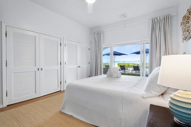 Bright coastal master bedroom with white quilted bed, louvered closet doors, and sliding glass doors opening to a patio with lounge chairs, blue umbrellas, and waterfront view.