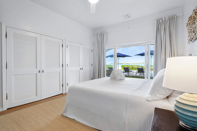 Bright coastal master bedroom with white quilted bed, louvered closet doors, and sliding glass doors opening to a patio with lounge chairs, blue umbrellas, and waterfront view.
