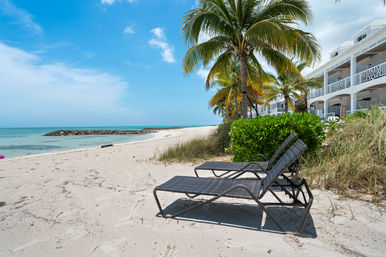 Two empty lounge chairs on a sandy tropical beach under swaying palm trees beside white beachfront villas, overlooking calm turquoise ocean and clear blue sky.