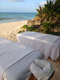 Oceanfront beach massage setup with two white-sheeted treatment tables on soft sand, turquoise sea, rocky shoreline and tropical greenery nearby.