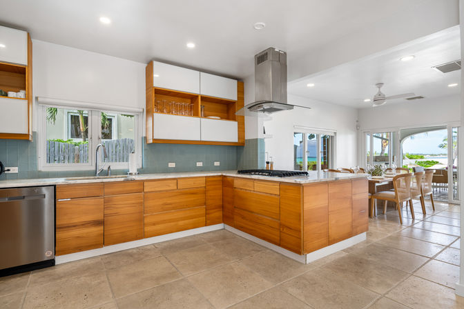 Bright open-plan coastal kitchen with warm wood cabinets, marble-topped island with gas cooktop and stainless steel range hood, light blue tile backsplash, stainless dishwasher, sunlit windows and sliding doors opening to a dining area and ocean view.