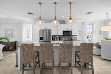 Airy modern open-concept kitchen with granite island, four woven rattan bar stools, glass pendant lights, stainless steel appliances, and white cabinetry.