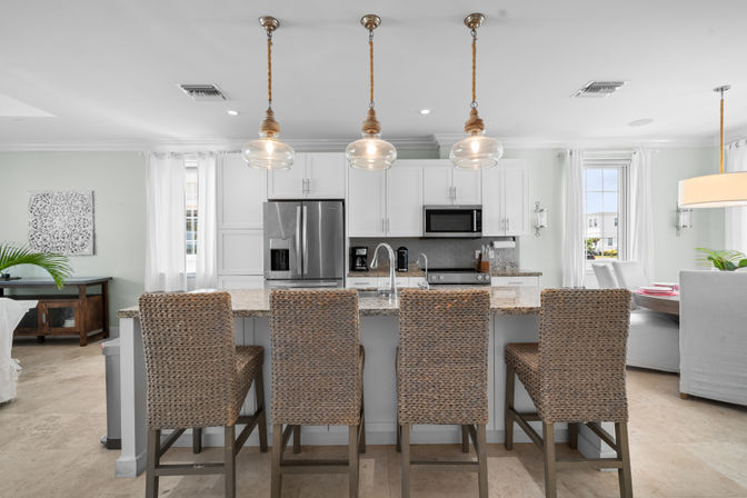 Airy modern open-concept kitchen with granite island, four woven rattan bar stools, glass pendant lights, stainless steel appliances, and white cabinetry.